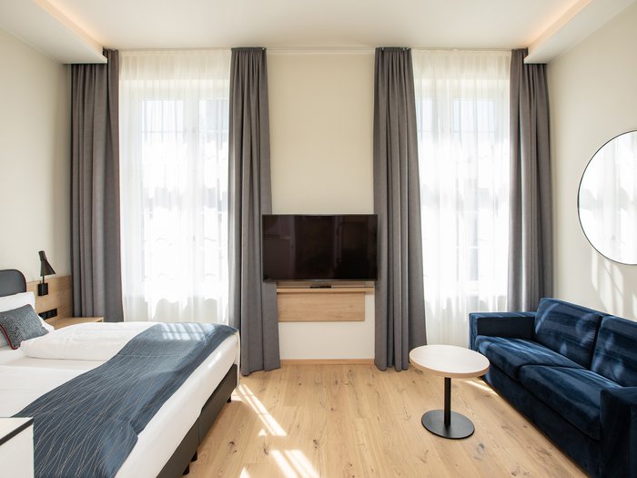 Hotel Böhlerstern in der Steiermark: unsere Zimmer Helles Hotelzimmer mit Doppelbett, blauem Sofa und Wandfernseher